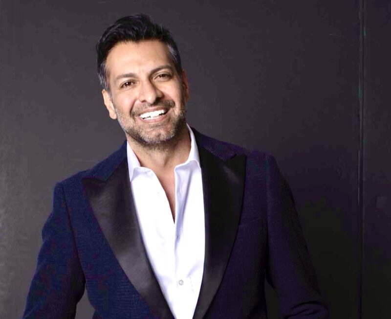 Smiling man in a dark suit with a white shirt, standing against a dark background.