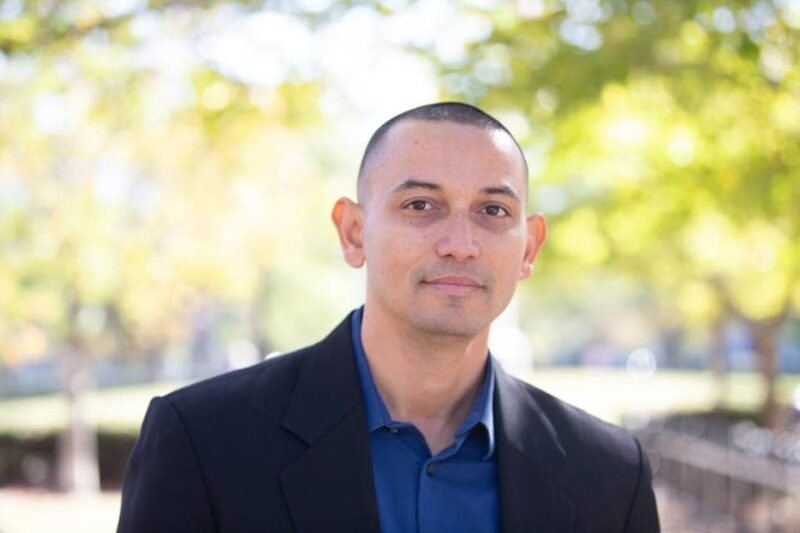 A person with a shaved head in a black suit and blue shirt stands outdoors with blurred trees and sunlight in the background.