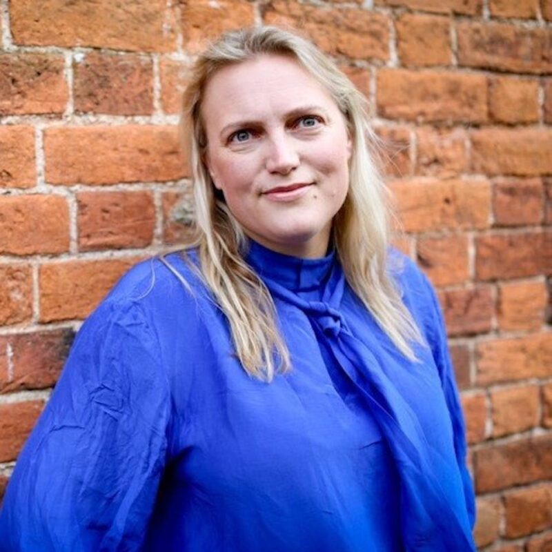 Person with long blonde hair in a blue blouse stands in front of a brick wall, smiling slightly.