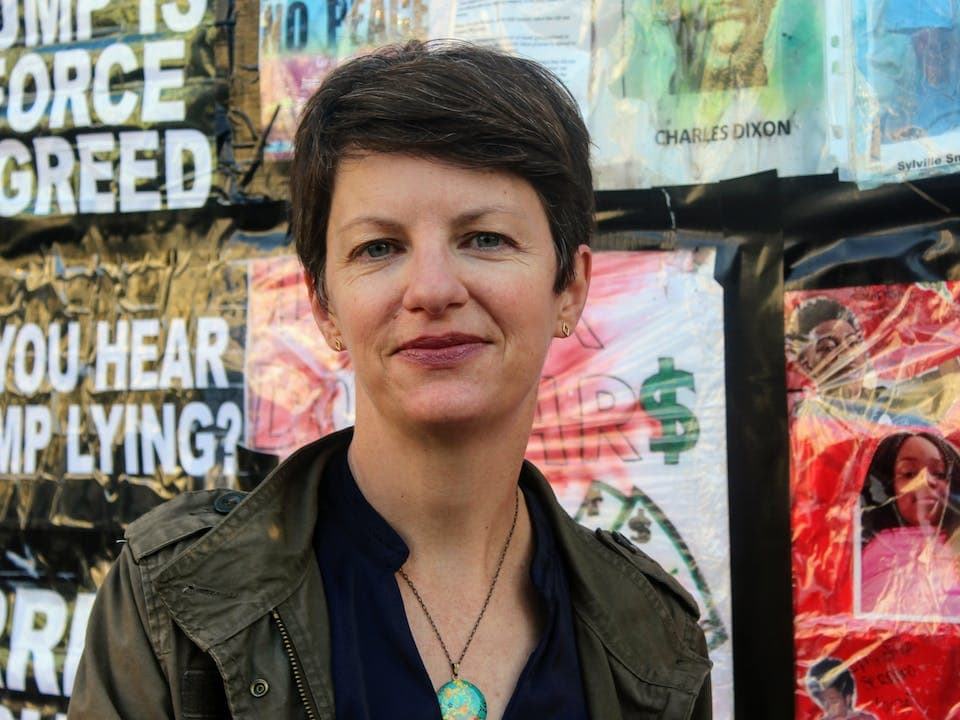 Person with short hair stands in front of posters and graffiti, wearing a dark jacket and necklace.