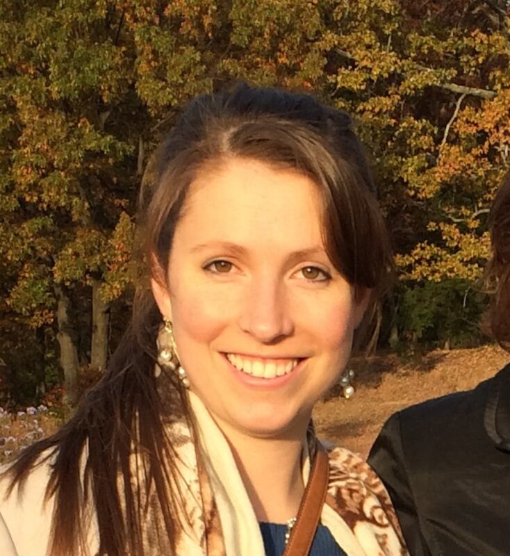 Woman smiling outdoors with autumn trees in the background.