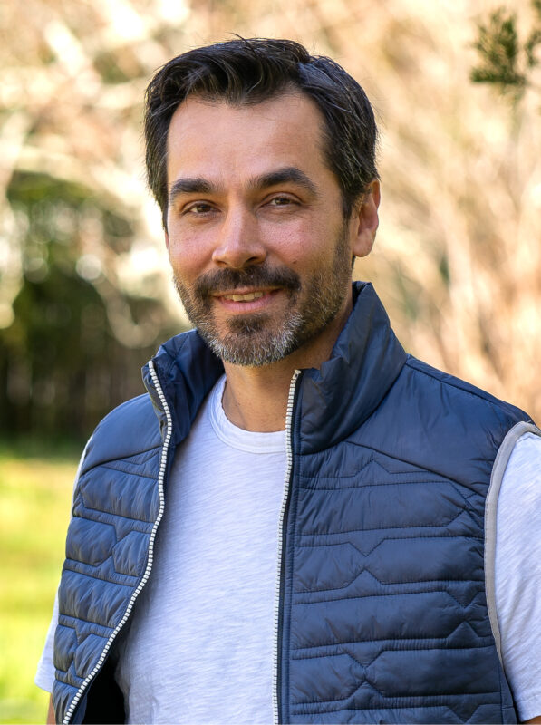 Man with a beard wearing a blue vest and white t-shirt, standing outdoors with blurred trees in the background, smiling at the camera.