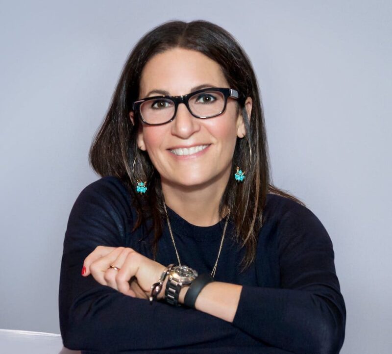 Smiling woman with glasses and shoulder-length hair, wearing a dark top and turquoise earrings, rests her arms on a surface.