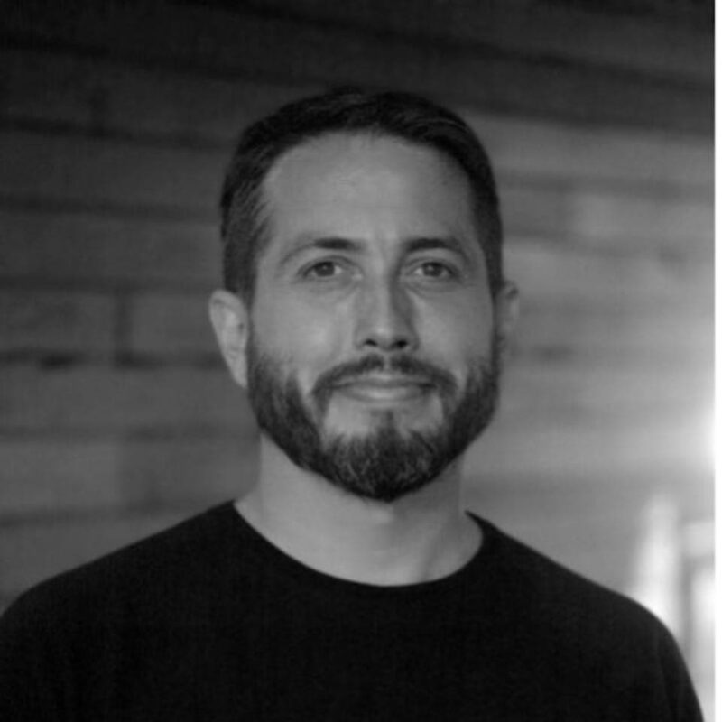 A man with short hair and a beard smiles, wearing a dark shirt, in front of a wooden wall. The image is in black and white.
