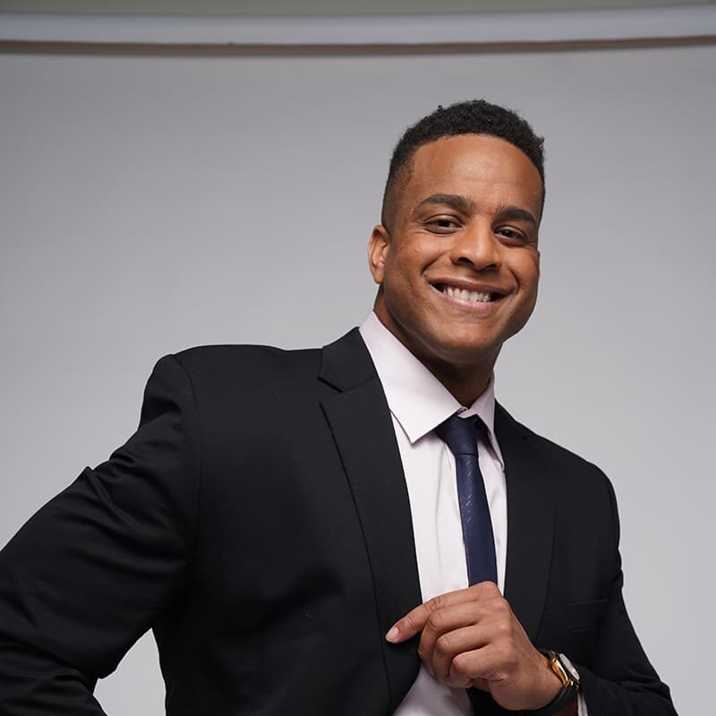Man in a black suit and tie smiles, standing against a plain background.
