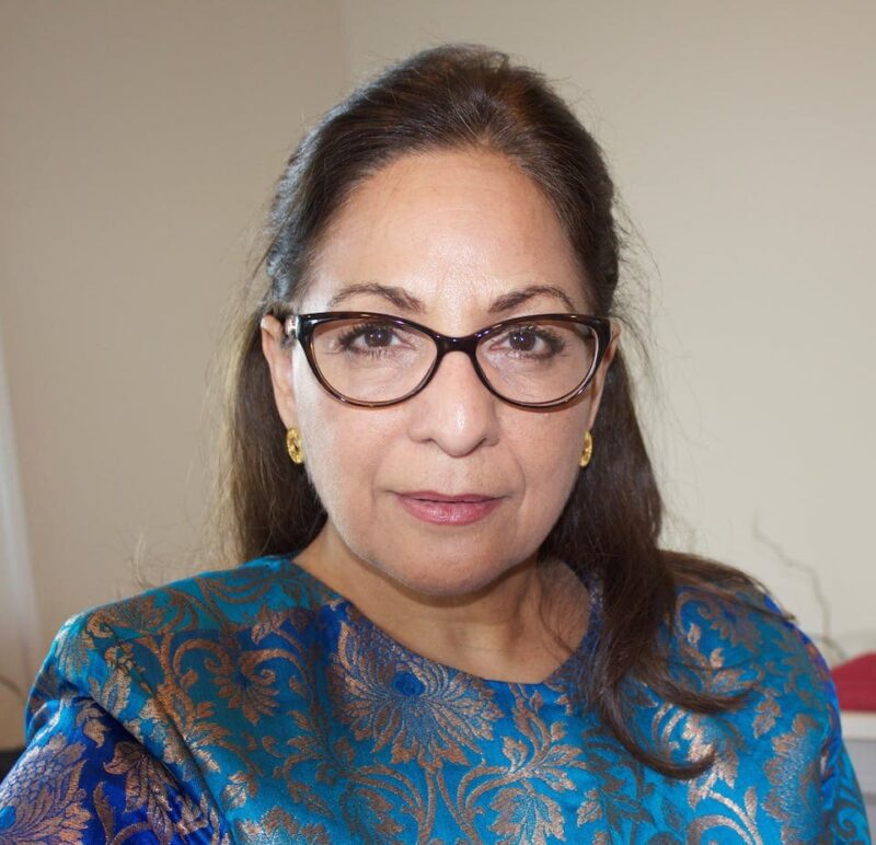 A person wearing glasses and a blue patterned top with gold accents, standing indoors.
