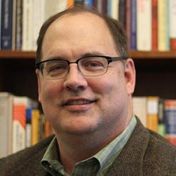 Man wearing glasses and a blazer smiles, standing in front of a bookshelf filled with books.
