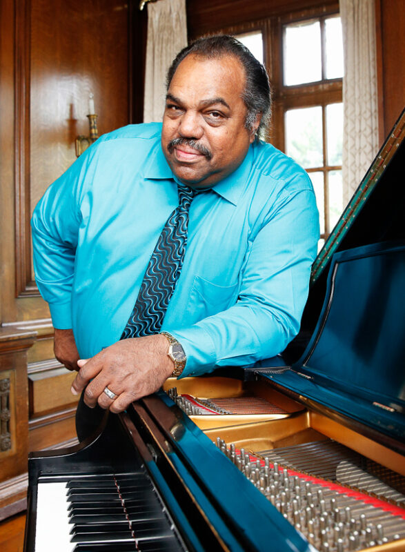 A person in a turquoise shirt leans on a piano inside a room with wooden walls and large windows.