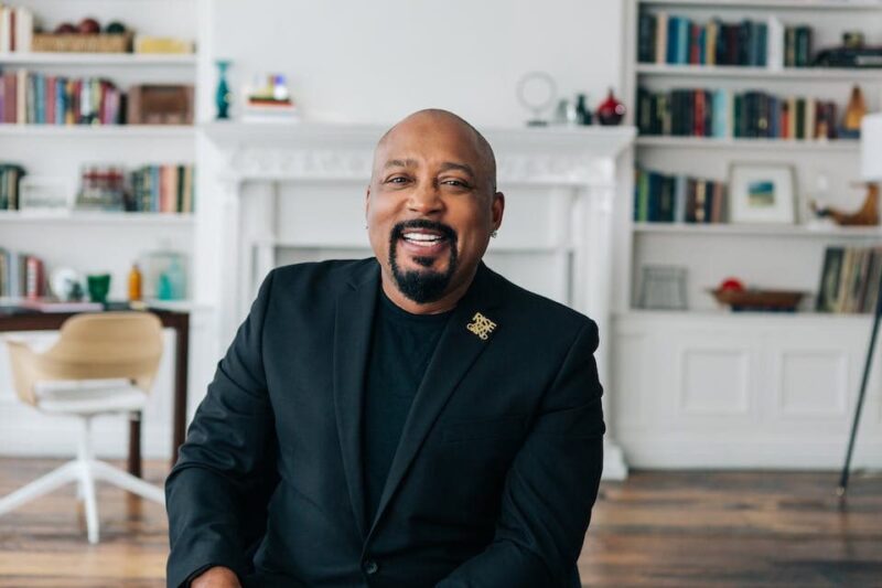 Man in a black suit smiling, sitting in a bright room with bookshelves and a fireplace in the background.