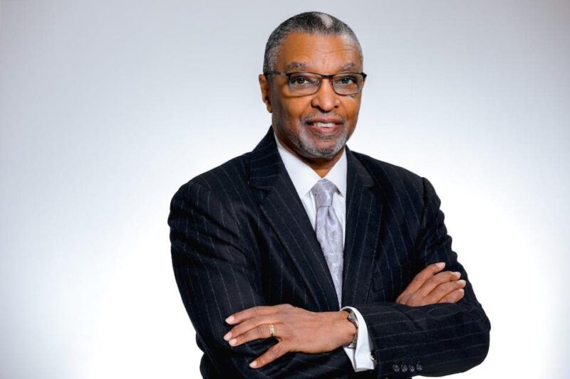 Smiling man in a pinstripe suit with glasses and a tie, arms crossed, against a light gray background.