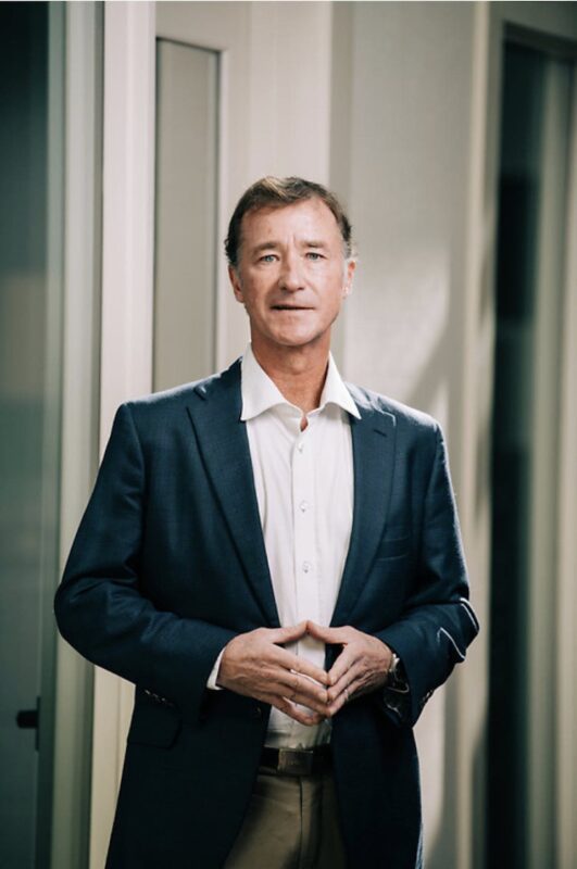 Man in a navy blazer and white shirt stands indoors with hands clasped, softly smiling.