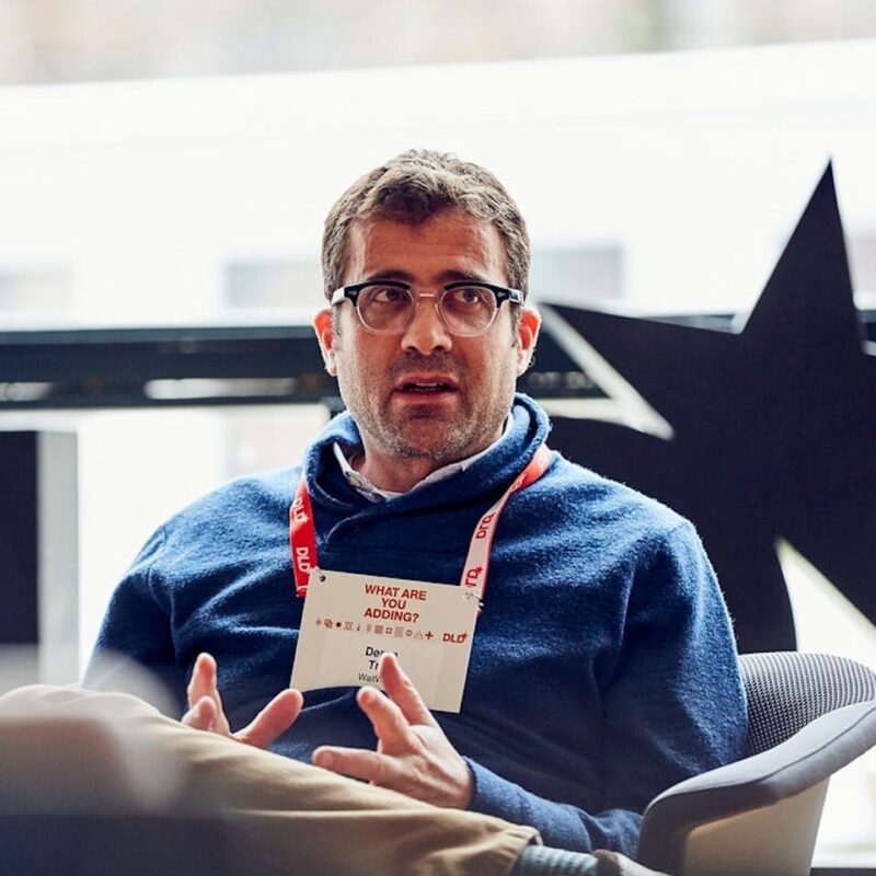 A person wearing glasses and a blue sweater speaks while seated indoors. They have a lanyard with a name tag.