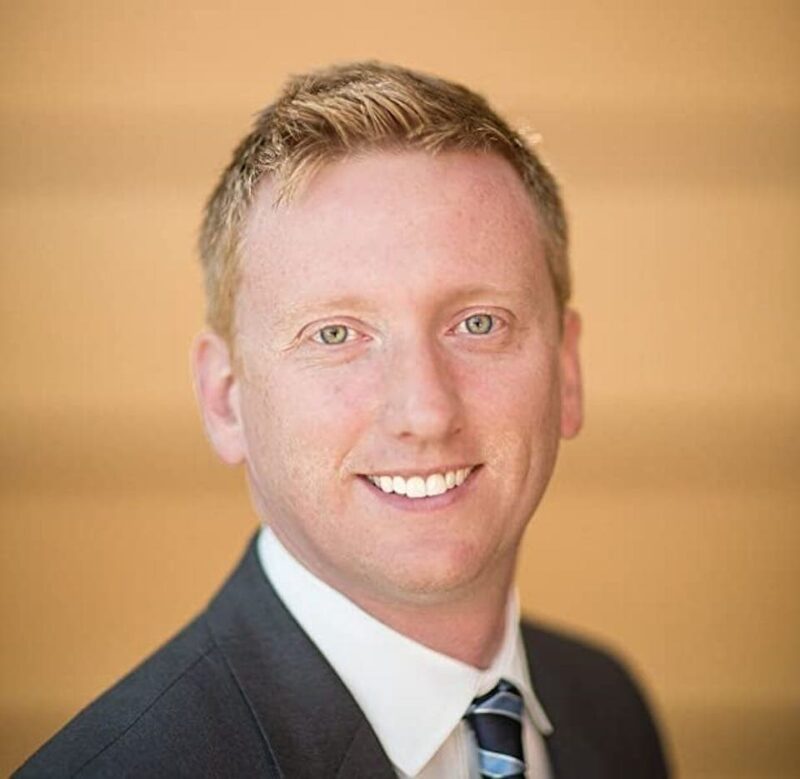Man in a suit and tie smiling against a blurred tan background.