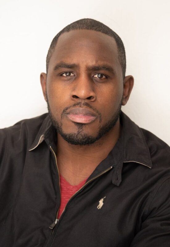 Man with short hair, beard, and serious expression wearing a black jacket over a red shirt, against a plain white background.