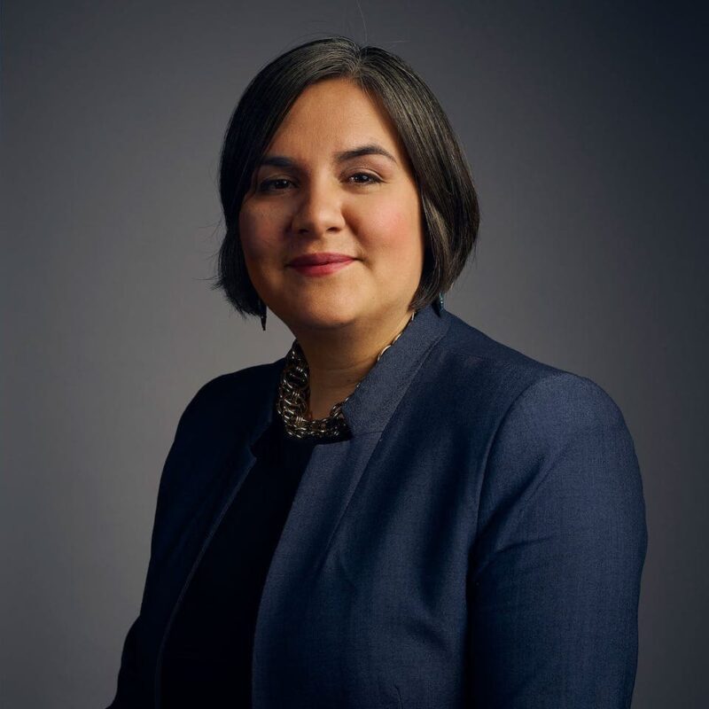 A person with short dark hair smiles slightly, wearing a dark blazer and necklace, set against a gray background.