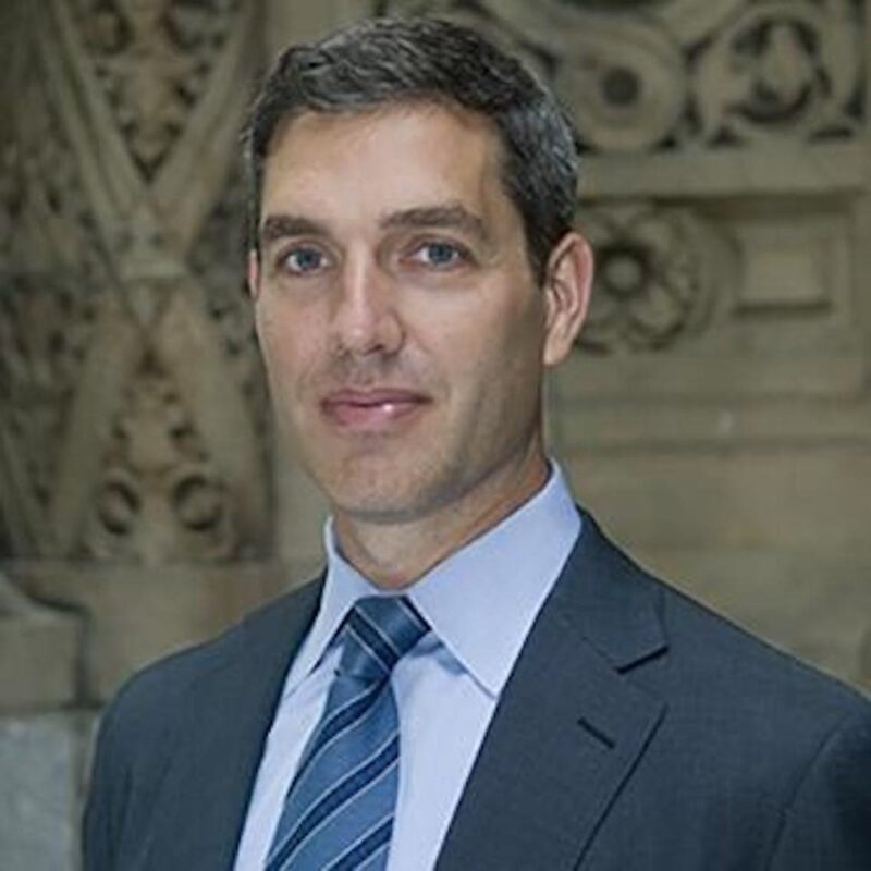 A man in a suit and striped tie stands in front of an ornate stone background.