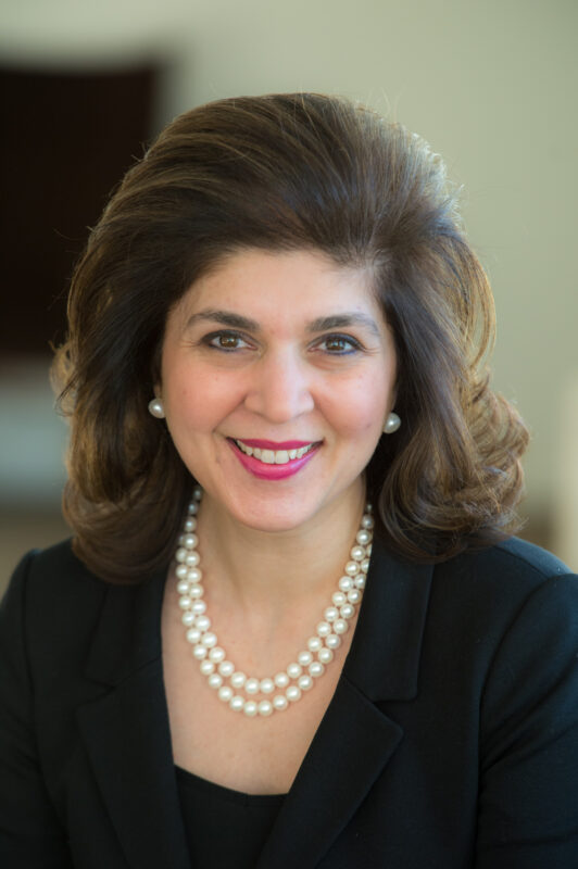 A woman with shoulder-length brown hair, wearing a black top and pearl necklace, smiles at the camera.