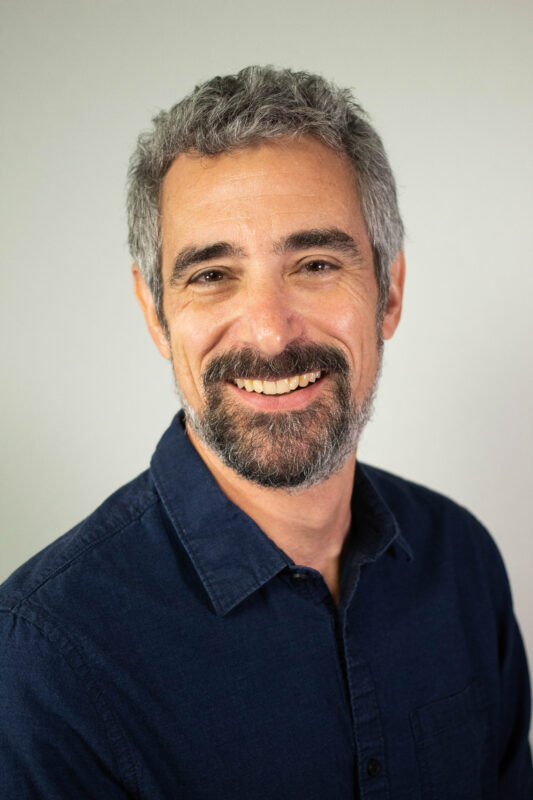 A man with gray hair and a beard smiles at the camera, wearing a dark blue shirt against a plain background.