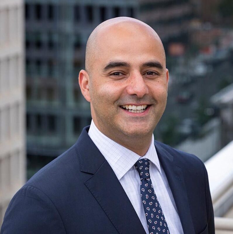 A bald man in a suit smiles at the camera, standing on a rooftop with an urban background.