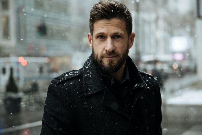 Man in a black coat stands outside in falling snow, with a cityscape background.