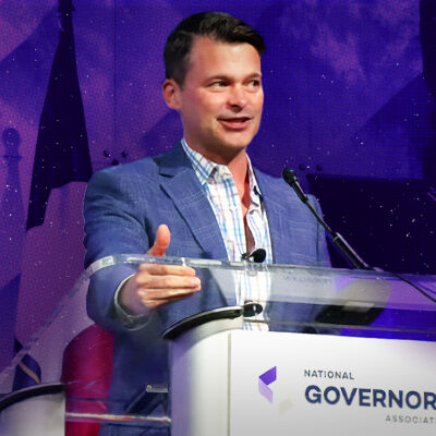 Man speaking at a podium with the National Governors Association logo, gesturing with his hand, against a background with stars and a flag.