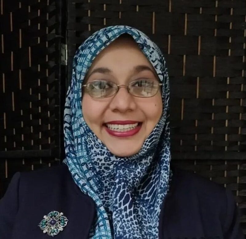 A person wearing glasses and a patterned hijab smiles broadly in front of a dark woven background.