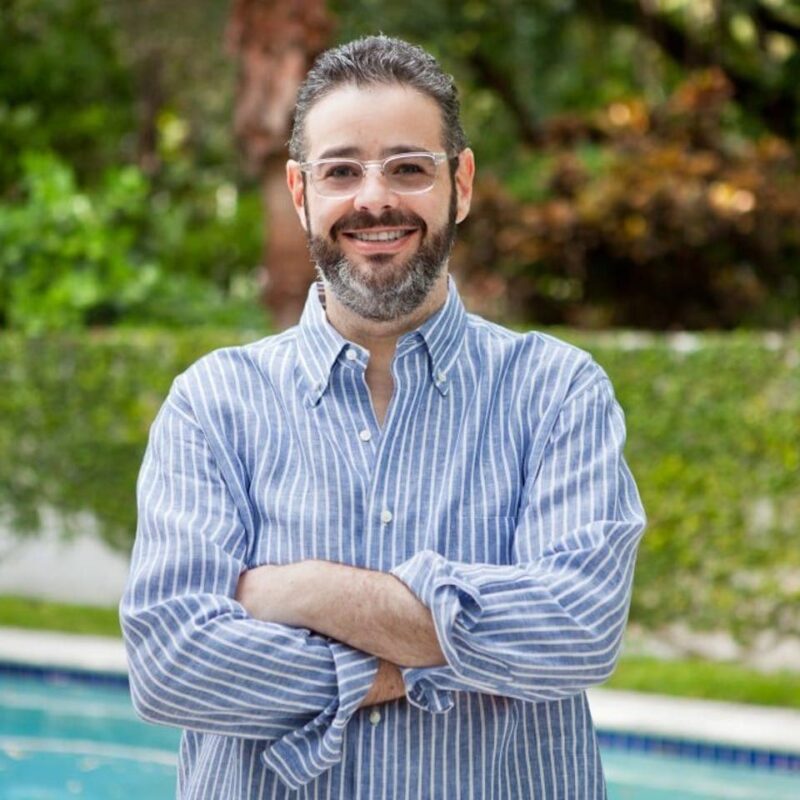 Person with glasses and beard smiling, wearing a striped shirt, stands with arms crossed in front of a pool and greenery.