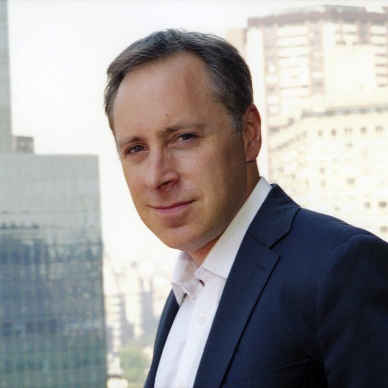 A person in a suit stands outdoors with a city skyline in the background.
