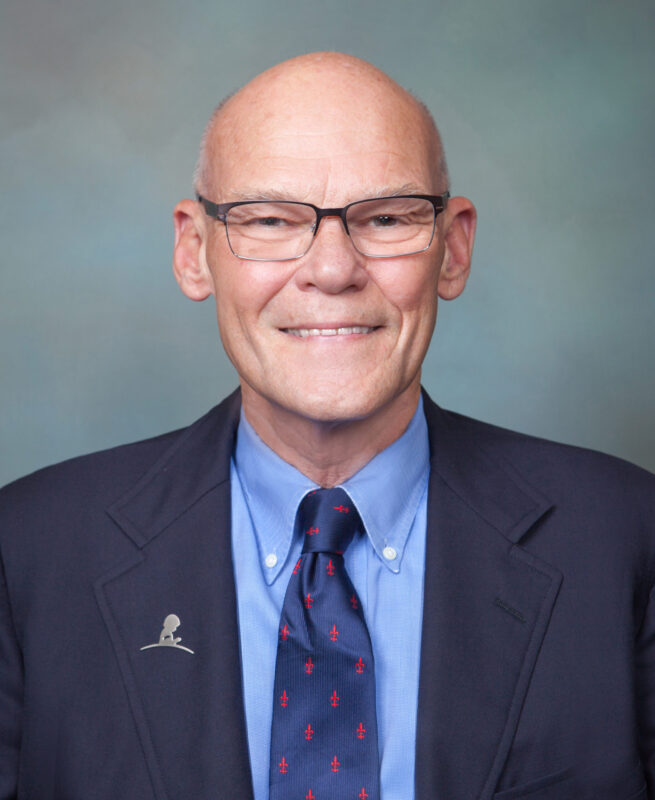 Bald man in glasses wearing a suit and tie with small red symbols, smiling at the camera against a plain background.