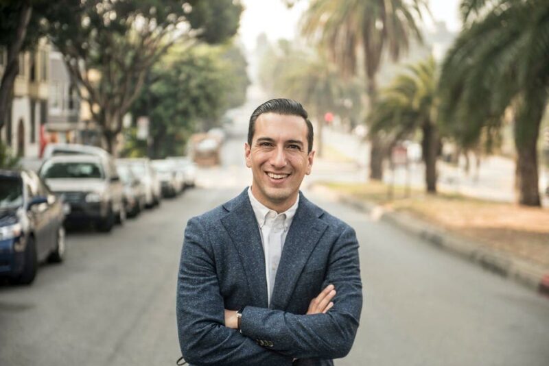 Smiling person in a blue blazer stands on a tree-lined street with crossed arms, surrounded by parked cars on a sunny day.