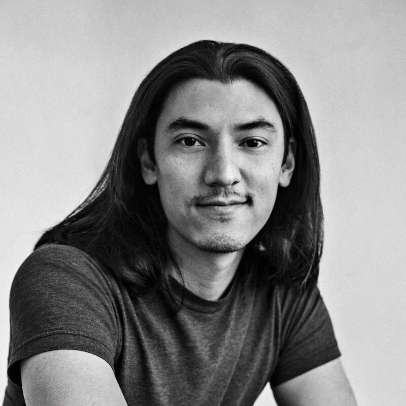 Monochrome image of a person with long hair, wearing a T-shirt, looking at the camera with a slight smile.