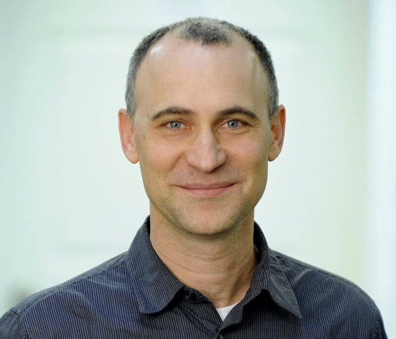 Man with short hair and a slight smile wearing a dark shirt against a light background.