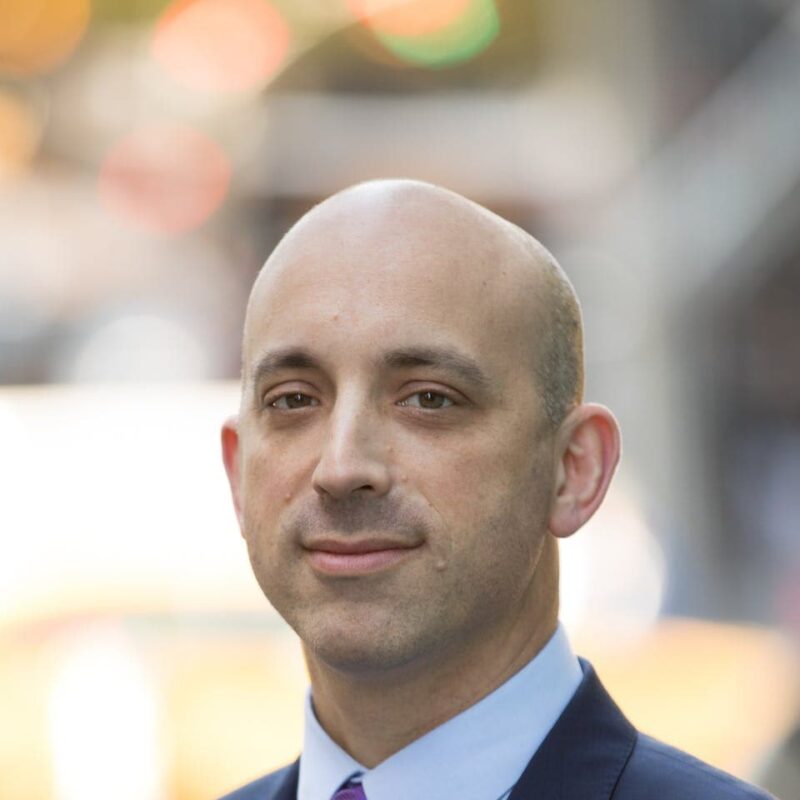 A bald man in a suit standing outdoors with blurred city lights in the background.