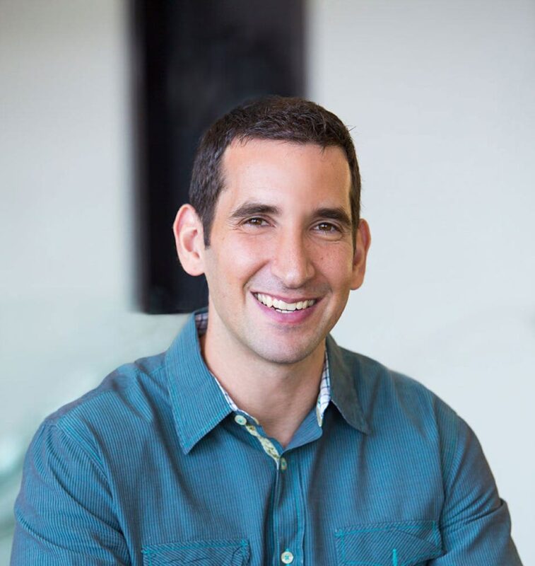 Smiling person with short dark hair in a teal shirt poses in front of a light, blurred background.