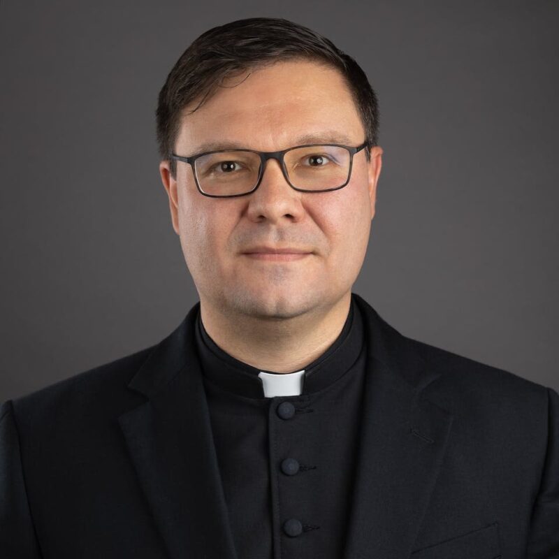 Man wearing glasses and a black clerical suit with a white collar, posing against a dark gray background.