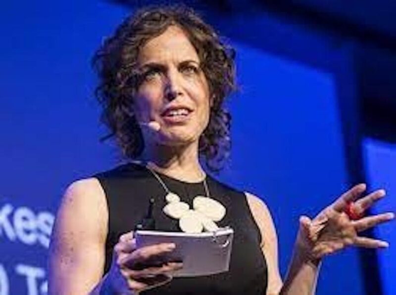 Woman with curly hair, wearing a black top and a large necklace, speaking on stage with a microphone and holding a notepad.