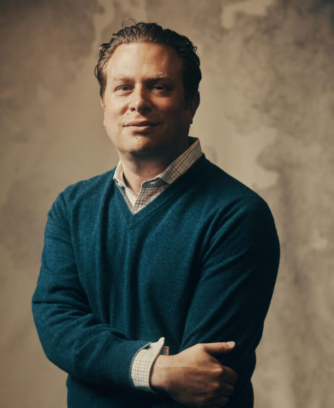 A person with short hair, wearing a blue sweater and a checked shirt, stands against a textured wall, looking confidently at the camera.