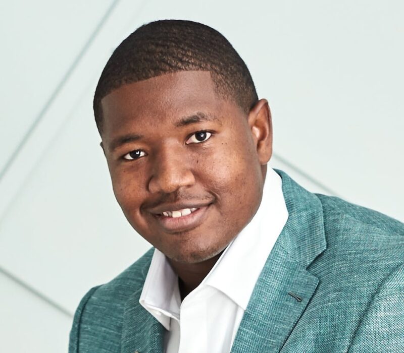 Smiling man in a teal blazer and white shirt against a light background.