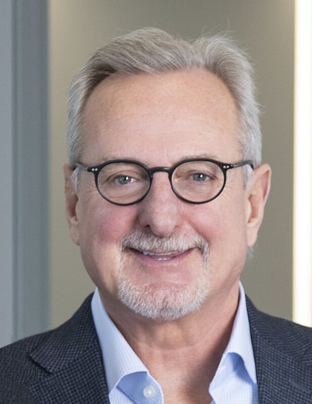 Man with glasses, a gray beard, and short gray hair, wearing a blue shirt and a dark blazer, smiles in a professional setting.