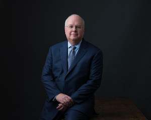 A man in a navy suit and tie sits on a wooden table against a dark background, hands resting on his lap.