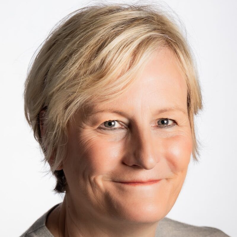 Smiling person with short, light hair and a light-colored top against a plain background.