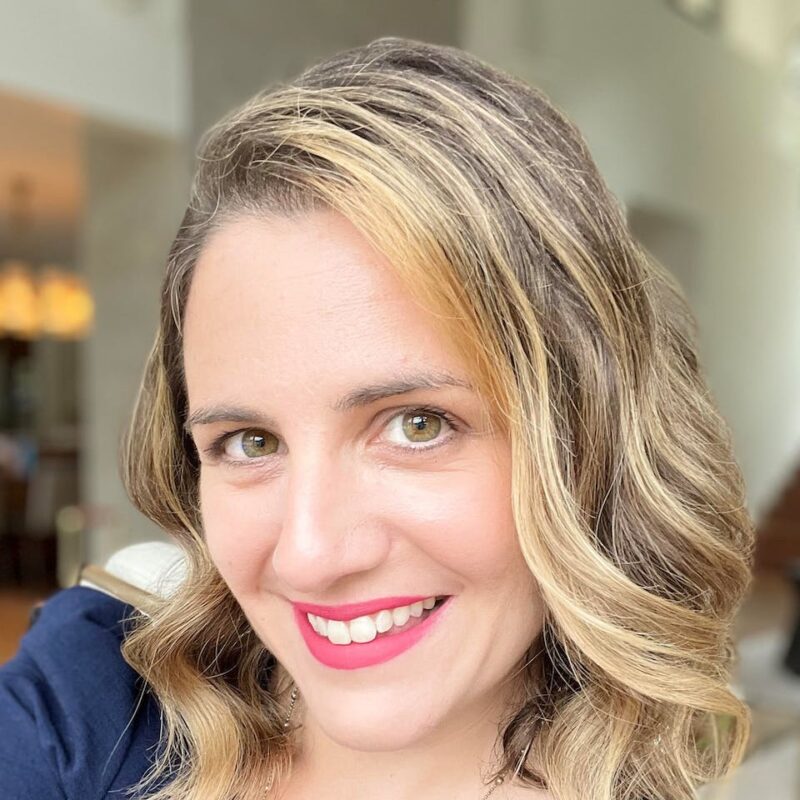 Person with wavy, shoulder-length hair smiling at the camera indoors. Soft lighting and blurred background with hints of furniture.