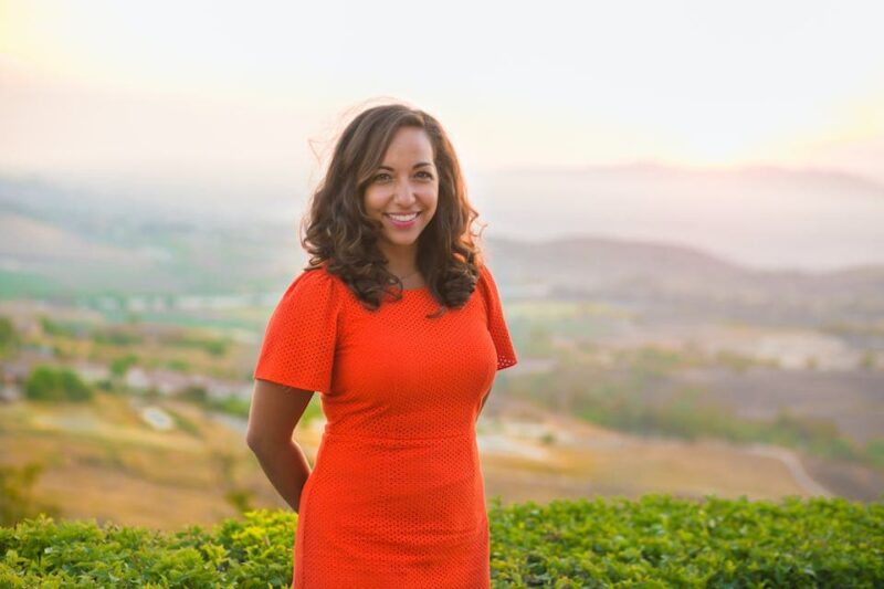 Smiling woman in an orange dress stands in front of a scenic landscape with rolling hills and a hazy sky.