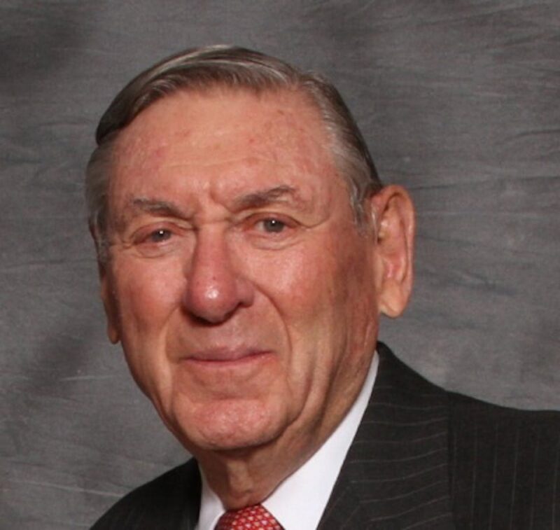 Elderly man in a suit and red tie against a gray backdrop, looking at the camera with a subtle smile.