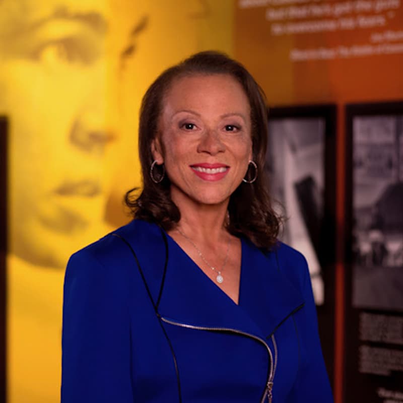 A woman in a blue blazer stands smiling in front of a yellow background with blurred images and text.