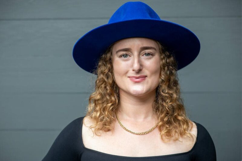 A person with curly hair wearing a blue hat, black top, and gold necklace smiles against a gray background.