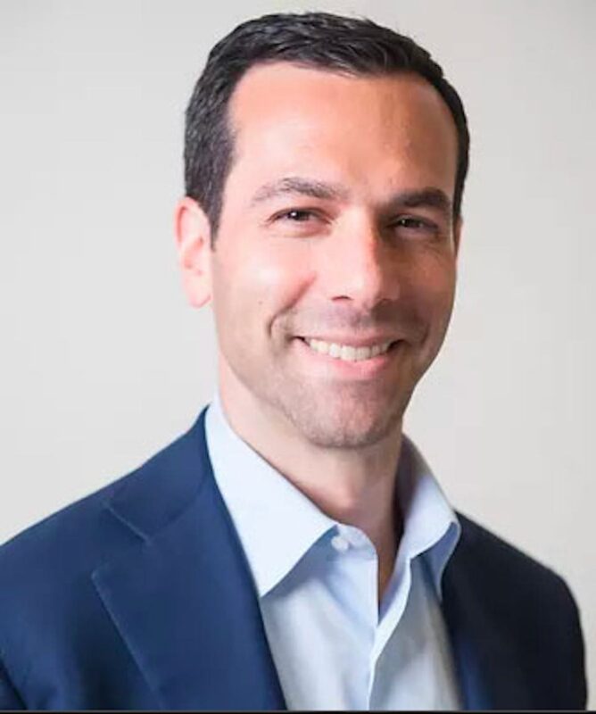 Smiling man in a blue suit and white shirt against a plain background.