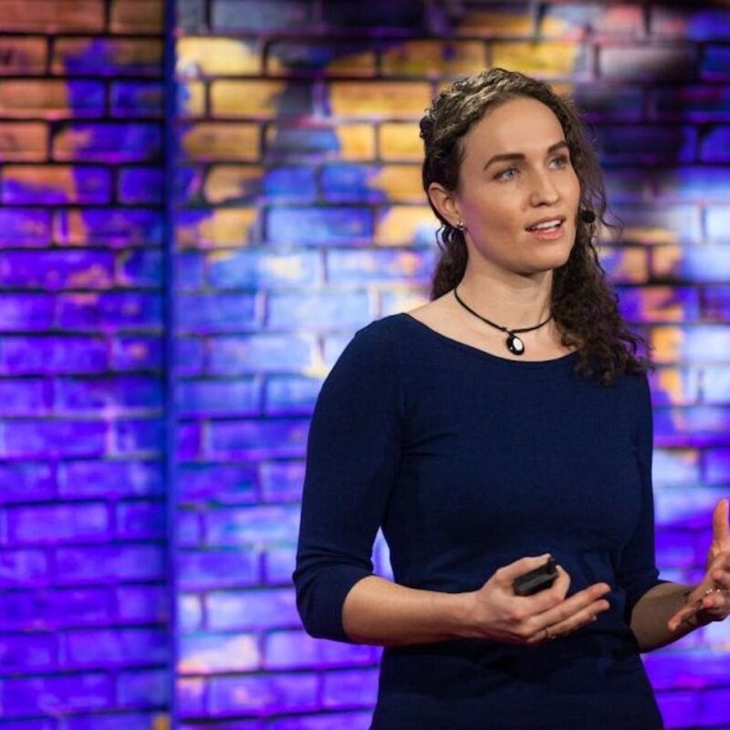 Person with curly hair speaking on stage against a colorful brick wall, holding a remote and gesturing with hand.