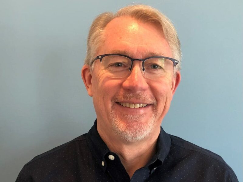 Man with glasses and a light beard smiling, wearing a dark shirt against a light blue background.