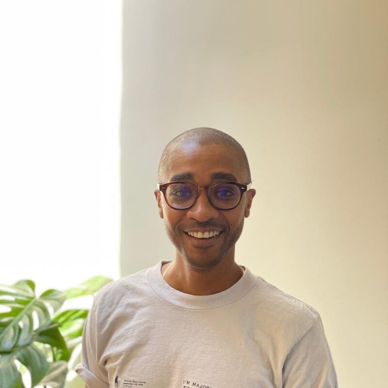 A person with glasses smiles indoors, wearing a light-colored shirt. A green leafy plant is partially visible on the left.